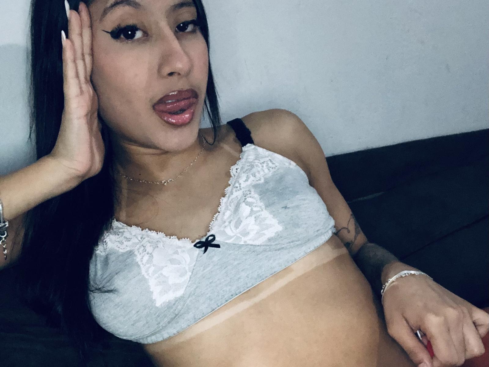 SantanaSuny with dark hair, wearing a lacey grey bralette, showing tan lines and puckered lips, hand near face, tattoo on right arm, bracelet on left wrist, sitting on black.