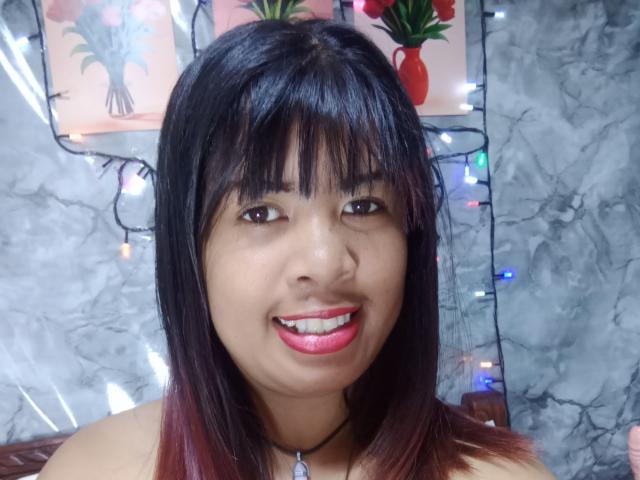 MariahSquirting69 has dark straight hair with bangs, brown eyes, and red lipstick. She's smiling with her pink-tinted tongue slightly visible. She's wearing a black necklace with a pendant.
