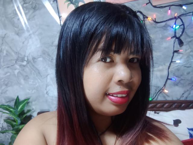 MariahSquirting69 with black and red-dyed straight hair, bangs, and red lipstick, smiling with slightly parted pink lips, showing her cute Asian face and medium brown skin.