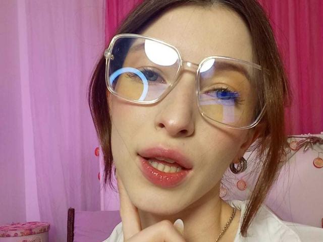 MeganSolar mit klaren Brillen, braunen Haaren, blauen Augen, einem Ringlicht, das im Auge reflektiert, rosafarbenen Lippen, einer Hand in der Nähe des Kinns, einem weißen Hemd, einem rosafarbenen und violetten Hintergrund, einer glänzenden Nase und einem leicht geöffneten Mund..