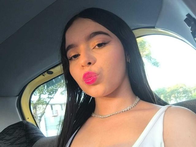 LissyCarter with long black hair, pink lipstick, and a silver necklace, making a pouty face in a car, wearing a white top, showing her tits slightly.
