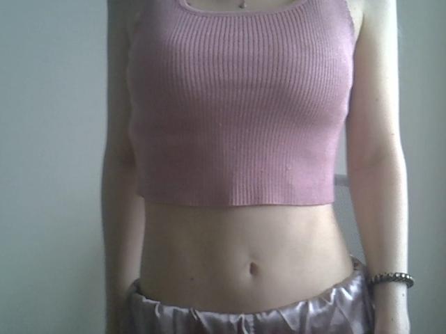 A close-up of DitaVira's upper body, showing a pink ribbed crop top that fits snugly, highlighting her small breasts. Her midsection is exposed, revealing a flat stomach with a visible navel. She wears satin-like, light purple pants with a slight ruffle at the waist. A simple bracelet is on her right wrist. The image is slightly blurry, emphasizing the casual, everyday style.