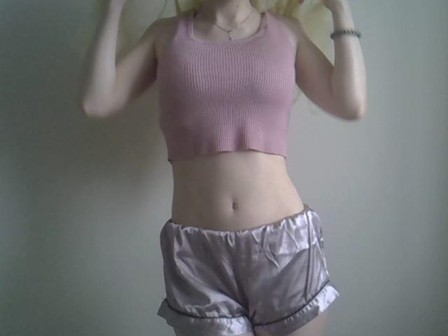DitaVira has pale skin, small tits, wearing a pink ribbed crop top and shiny purple shorts with a ruffled bottom, hands raised, a thin necklace, and a bracelet.