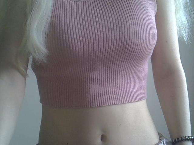 Close-up of DitaVira's torso showing a pink ribbed crop top. Her blonde hair is partially visible on the left. The top is tight, highlighting her small breasts. She has a fair skin tone with a subtle navel. Her right arm is slightly raised, and she wears a black bracelet on her wrist. The image focuses on her midsection, with a slight shadow on her right side. The texture of the top is ribbed, and the color is a muted pink.