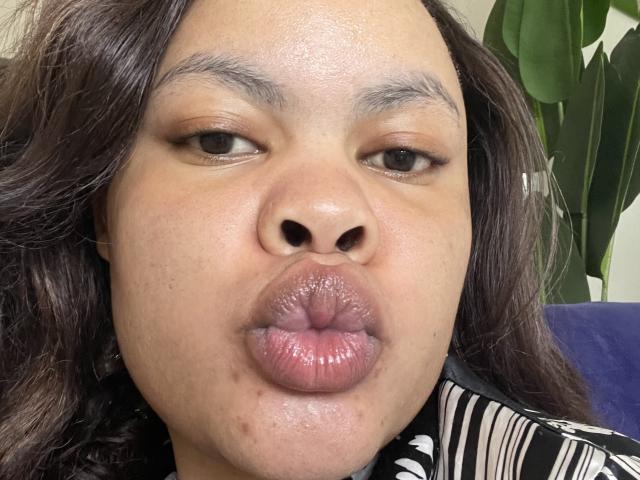 LucindaMartie's face with glossy, puckered lips making a kissy pout. She has dark, wavy hair, light brown skin, and subtle makeup. Her nose is small with a slight bump.