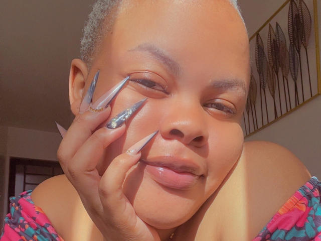 LucindaMartie with short gray hair, smiling, holding her cheek with long, silver, glittery nails, wearing a colorful patterned top, sunlight on her face, slightly glossy lips.