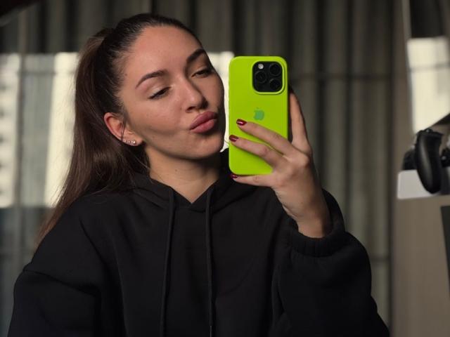 AquaLady with dark brown hair in a ponytail, wearing a black hoodie, taking a selfie with a neon green iPhone, pink nail polish, and a cute facial expression.