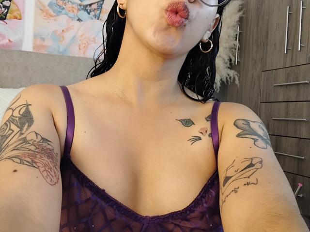 Mllevv with puffy pink lips, wearing a purple sheer top, showing tattoos on arms, wearing hoop earrings, and wet black hair, taking a selfie. Boobs, tattoos, and lips.