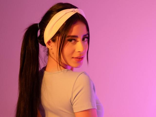 MadissonBlue met een witte hoofdband, een lange zwarte paardenstaart en een lichtgrijs shirt, kijkt over haar schouder, met roze en paarse verlichting. Ze heeft schattige oorbellen en een lichte glimlach..