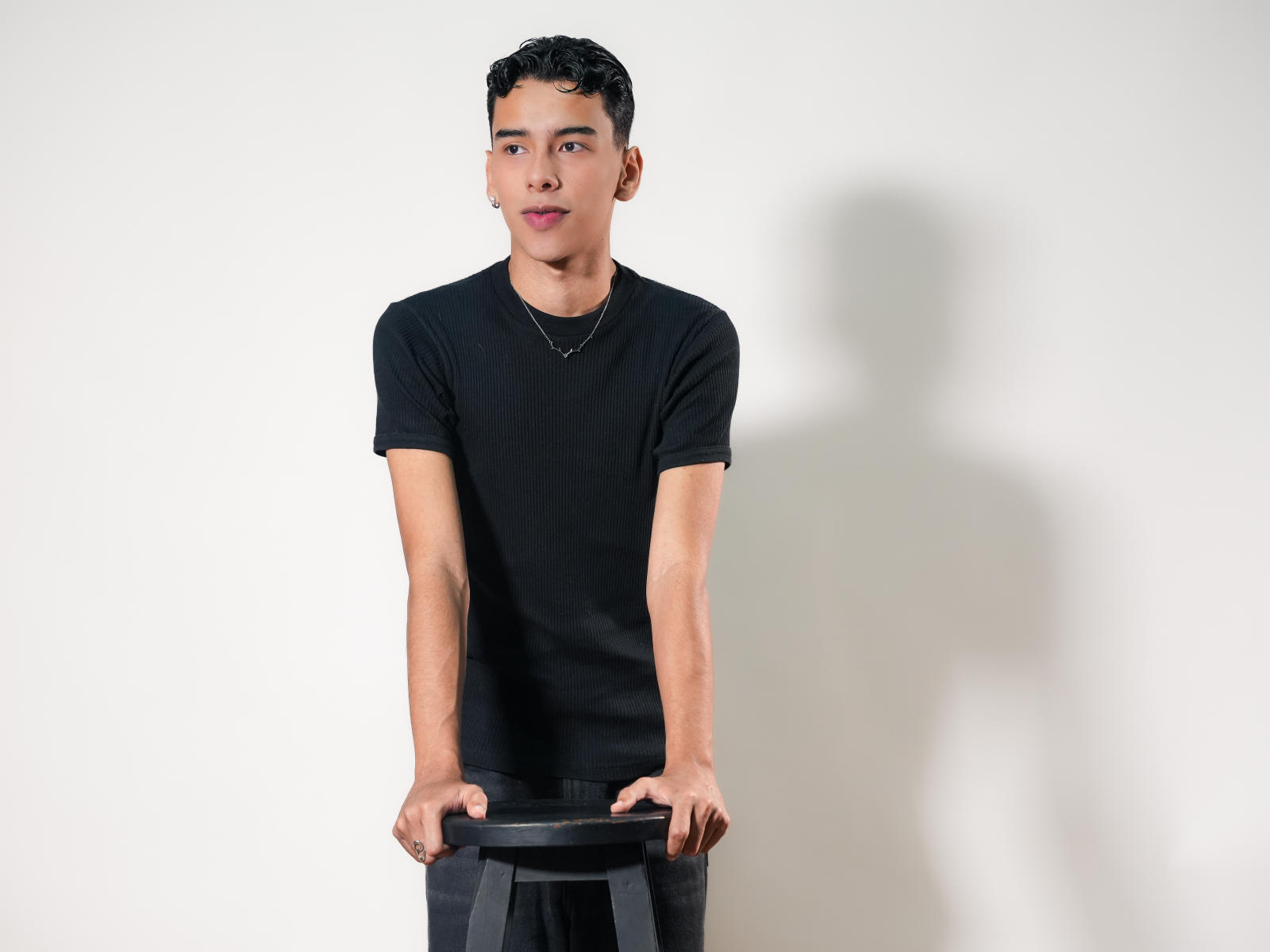 Aiden Royce mit kurzen, schwarzen, lockigen Haaren, der ein schwarzes T-Shirt und graue Hosen trägt, steht und hält einen schwarzen Hocker, ein Schatten fällt auf die weiße Wand, er schaut in die Kamera..
