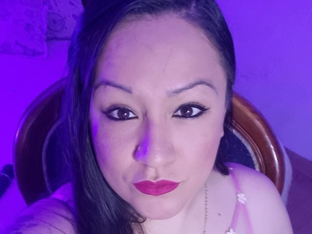 Close-up of LucianaDia with medium skin tone, dark straight hair, and purple lighting. She has thick black eyebrows, large almond-shaped brown eyes with black eyeliner, and red lipstick. Her left arm is slightly visible with a pink strap, and she's wearing a thin gold necklace. LucianaDia's face is centered, looking directly at the camera with a slight smile. The background is blurred, focusing on her face. Her hair is parted to the left.