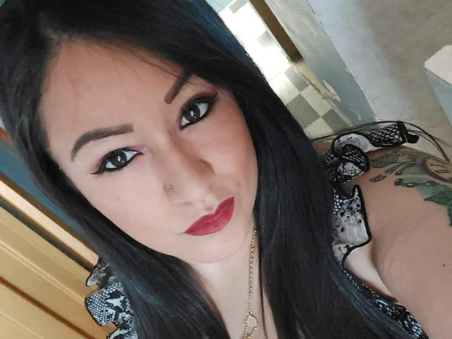 Luciana Dia ha lunghi capelli neri, sopracciglia scure e rossetto rosso. Indossa una maglietta a righe nere e bianche e ha dei tatuaggi visibili sulla spalla. Sta guardando la fotocamera..