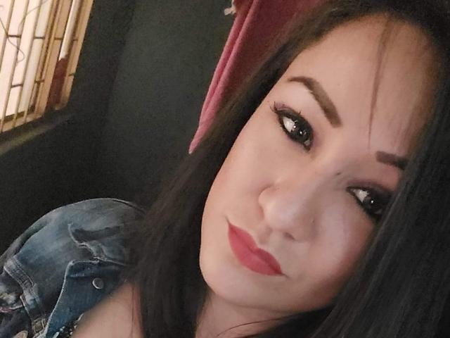 Close-up photo of LucianaDia with dark brown hair, slightly wavy, and thick eyebrows. She has light skin, dark eyeliner, and red lipstick. LucianaDia is looking to the side with a subtle smile. She wears a denim jacket with a slightly textured fabric. A pink towel is partially visible in the background. The light source is from the left, creating a soft shadow on her right side. Her makeup is bold and defined.