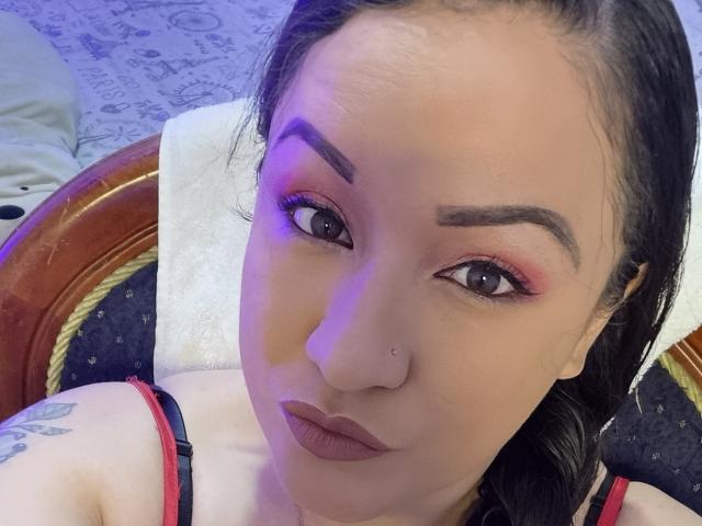 Close-up of LucianaDia with light brown skin, dark eyebrows, and red eye makeup. She has a small nose piercing on the right side and is wearing dark brown lipstick. LucianaDia's black hair is partially visible. She is seated on a chair with a wooden frame and a white towel behind her. She has a tattoo on her right shoulder. The image has a blue light on her left cheek. The chair has a dark, patterned fabric. Her expression is neutral.