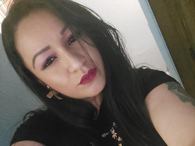 LucianaDia has long black hair, purple eyeshadow, and dark red lipstick. She wears a black top with floral patterns. Her right arm shows a green tattoo. She has a small nose piercing on her left side. Her eyebrows are thick and dark. She looks slightly to the right with a neutral expression. The image has a casual selfie style. She wears small triangular earrings. The lighting is soft.