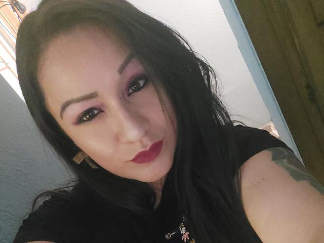 Luciana Dia a de longs cheveux noirs, du fard à paupières rose, du rouge à lèvres rouge et un tatouage sur son bras droit. Elle porte une chemise noire et a une expression sérieuse, avec un léger sourire..