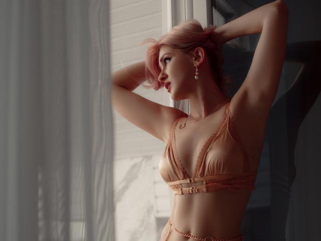 Lustarela with pink hair, arms raised, wearing a tan lace bra, showing her small tits, slim, pale skin, near a window with sheer curtains. Her earrings catch light.