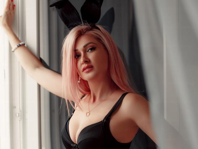 Lustarela with pink hair, black bunny ears, and a black bra, showing off her big tits and a cute face, standing by a window with her arm raised.