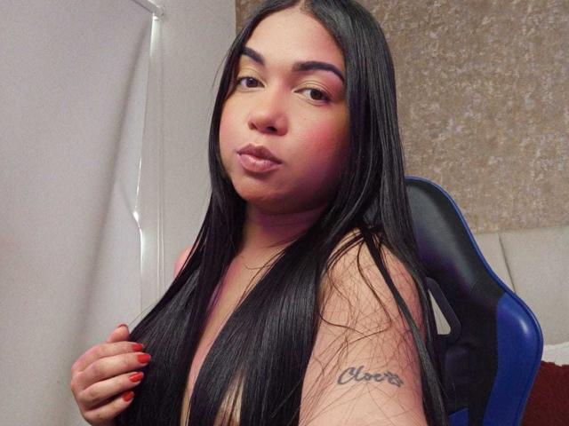 AliceSmity with long black hair, pinkish-red lipstick, and red nails, pouting with a tattoo on her right arm that reads "Cher". She's sitting in a blue chair.