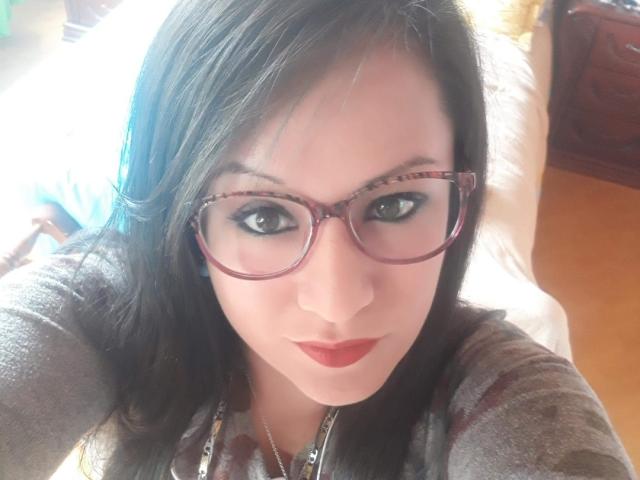 Close-up selfie of LucianaDia with medium-dark skin, black hair, and brown eyes. She wears red-tinted glasses and has dark red lipstick. Her eyebrows are black and slightly arched. She's wearing a gray, patterned shirt with a silver necklace. The image has bright, natural light from the left, highlighting her face. The background is blurred, focusing on her facial features. The texture of her hair is smooth, and her expression is neutral.