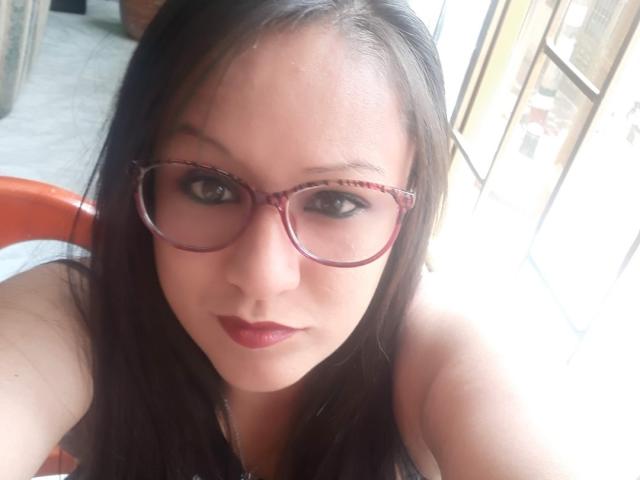 LucianaDia with dark brown hair, wearing red glasses and red lipstick, looking at the camera, showing her bare shoulders and a slight smile. Her tits are partially visible.