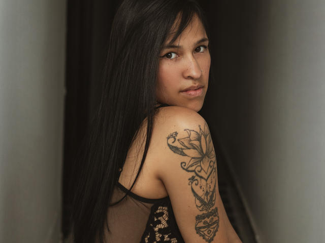 Andrea Brunert, com seus longos cabelos pretos, olhando por cima do ombro, tem uma tatuagem grande no ombro direito, com um design floral detalhado, e está vestindo uma blusa preta de renda..