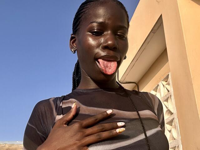 Close-up of KharlaQueen, dark-skinned with braided hair, sticking out her tongue. She wears a sheer black top with geometric patterns. Her right hand, with white and yellow-painted nails, rests on her chest. The sun casts sharp shadows on her face and top. She has small earrings in both ears. The image is sharp, with a clear blue sky in the background. Her skin has a glossy, shiny texture.