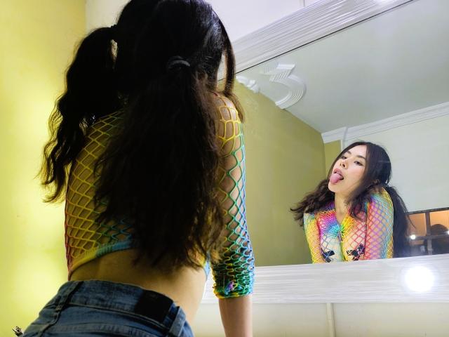 AliceEvansLove with long dark hair in pigtails, fishnet yellow top, tongue out, looking in mirror, reflection shows her face, wearing denim shorts, mirror has white frame, she's cute.