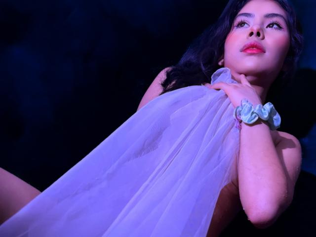 AliceEvansLove with purple-tinged skin, dark wavy hair, and pink lipstick, holding a sheer white cloth over her tits, wearing a blue scrunchie, against a dark background.