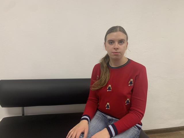 MaryCholls est une jeune fille mignonne avec un visage sérieux, portant un pull rouge avec de petits motifs triangulaires et un jean bleu, assise sur un banc noir..