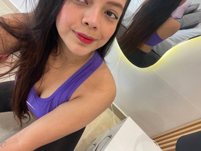 ViolettaRouse with long black hair, red lipstick, purple sports bra, and black pants, taking a selfie, showing her cute face and small boobs in a mirror.