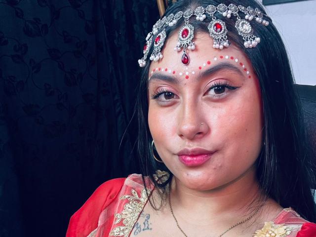 KalpanaAisha with dark hair, red eyes, and pink lipstick, wearing a jeweled headpiece with red gems and pearls, red and gold dress, small nose ring, and hoop earrings.