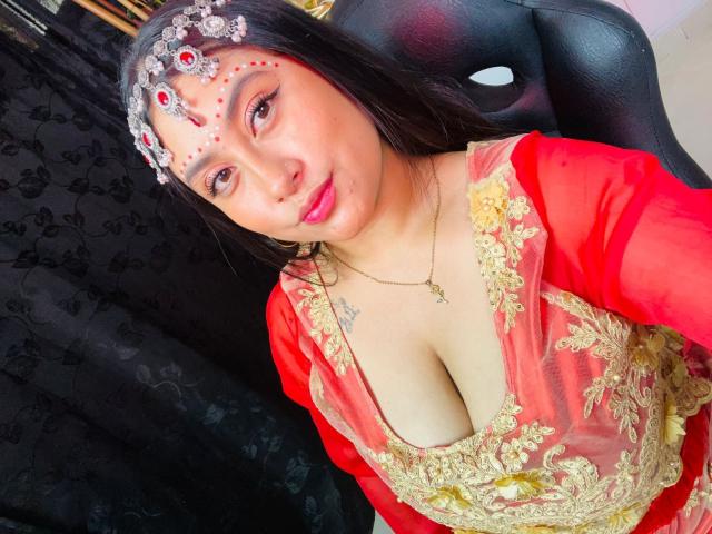 KalpanaAisha with dark hair, red and gold dress, cleavage, red face paint, headpiece, necklace, sitting on black chair, looking at camera, slight smile, light skin, small tattoos on chest.