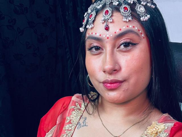 KalpanaAisha with dark hair, red lipstick, and a jeweled headpiece, has red and white facial markings, wearing a red and gold embroidered top, showing a small chest tattoo.