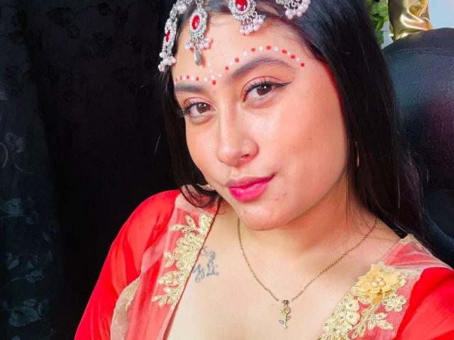 KalpanaAisha with dark hair, red makeup, and a headpiece, wearing a red and gold dress, showing her tits, nose ring, and tattoo, with pink lipstick and a necklace.