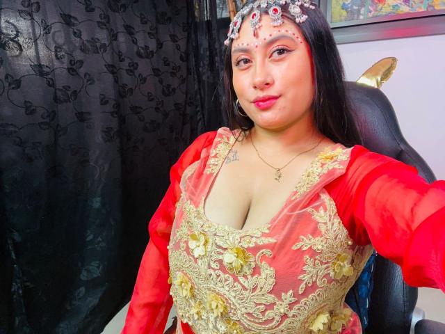 KalpanaAisha with big tits in a red and gold embroidered dress, red sleeves, jeweled headpiece, red lipstick, black curtain, and a chair in a brightly lit room.