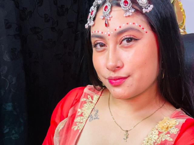 KalpanaAisha with dark hair, red lipstick, nose ring, and jeweled forehead piece, wearing a red and gold traditional outfit, showing small tits and a tattoo under her chest.