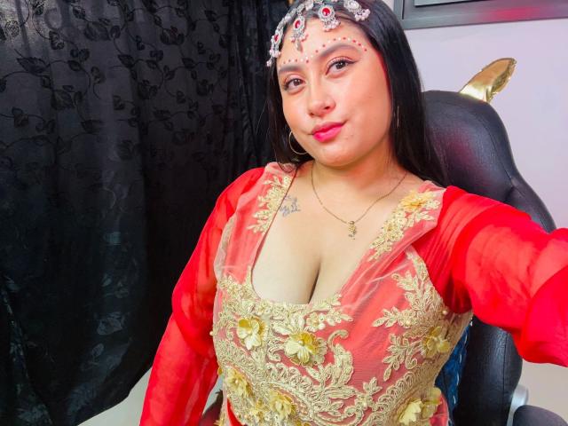 KalpanaAisha with medium tits in a red and gold embroidered dress, wearing a floral headpiece, sitting in a black chair, smiling at the camera.