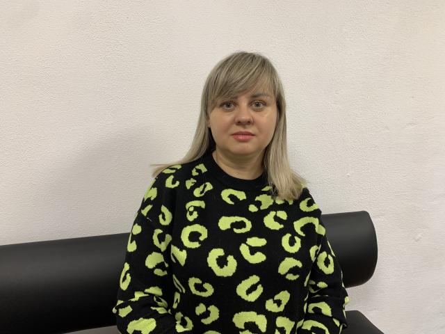 VeronicaSvitt with blonde hair, wearing a black leopard print sweater, sitting on a black bench, looking forward with a neutral expression.