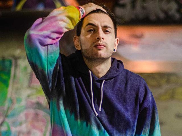 NachoMasterVidal with short brown hair, wearing a tie-dye hoodie and blue ear gauges, has a serious look, hand in hair, standing in a graffiti-laden urban setting.