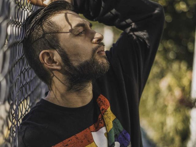NachoMasterVidal with a beard, black shirt, and colorful graphic, leaning against a chain-link fence, sunlight casting shadow on his face, short brown hair, raised arm, looking side.
