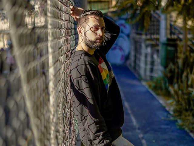 NachoMasterVidal leans against a chain-link fence, arms up, wearing a black shirt with a colorful diamond pattern, sunlight casting shadows, short beard, looking serious, urban background.