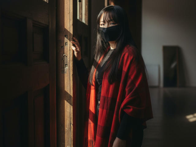BeloverNana with long black hair, wearing a red plaid shawl, black mask, and black top, standing in a wooden doorway, sunlight casting shadows on her face.