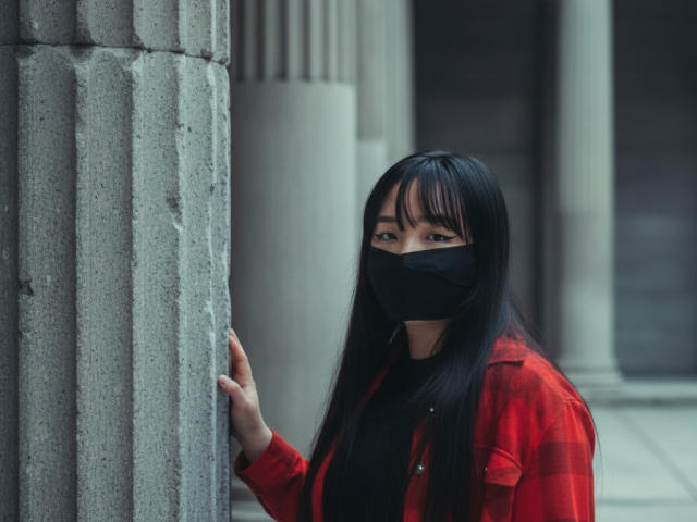 BeloverNana stands beside a gray stone column with vertical fluting. She has long black hair with bangs, wears a red plaid shirt over a black top, and a black face mask. Her right hand rests on the column. The image has a muted, urban feel with a focus on the contrast between her red shirt and the gray stone. Her expression is neutral.