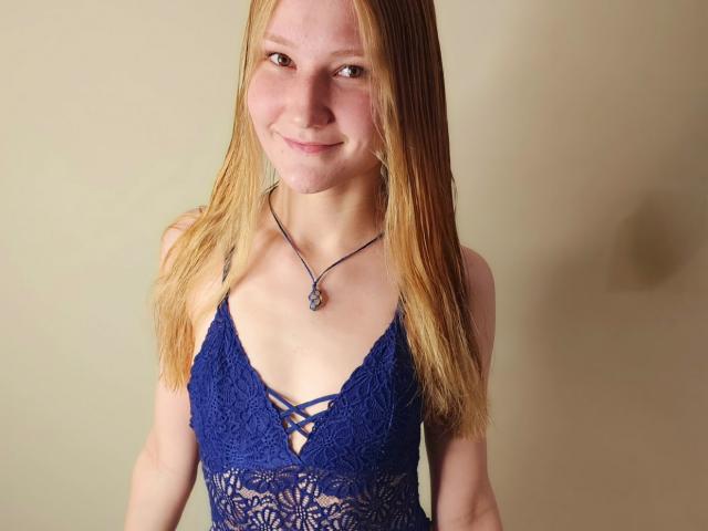 VanessaNorris with blonde hair, smiling, wearing a blue lace dress with a crisscross pattern over her small tits, and a black necklace with a blue stone.
