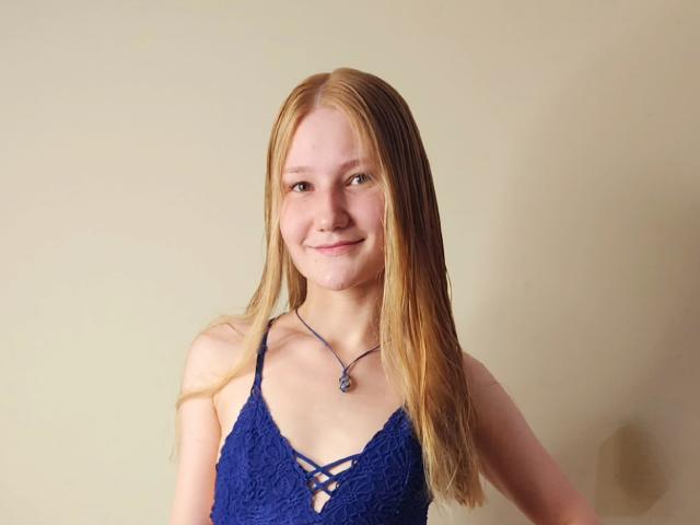 VanessaNorris with long blonde hair, blue lace top, small tits, smiling, wearing a blue necklace with a purple pendant, against a plain beige background.