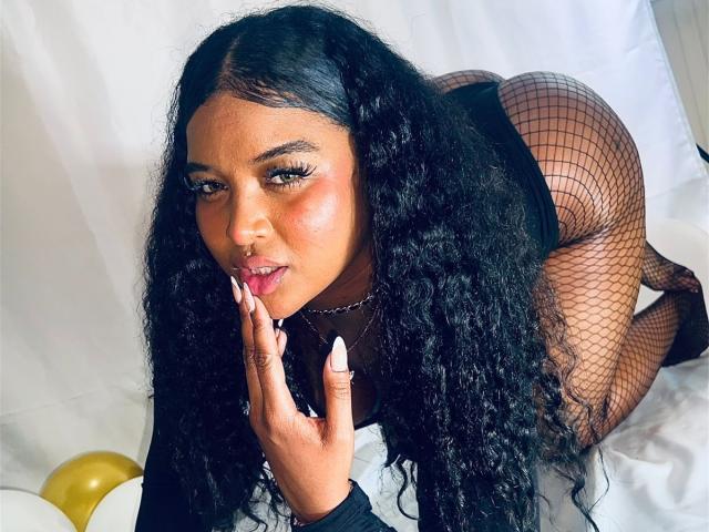 ChloeeFoxySs com cabelos pretos longos e cacheados, usando meia-calça de rede, blusa preta e unhas brancas, ajoelhada, com a mão no rosto, olhando para a câmera, mostrando seu bumbum arredondado..