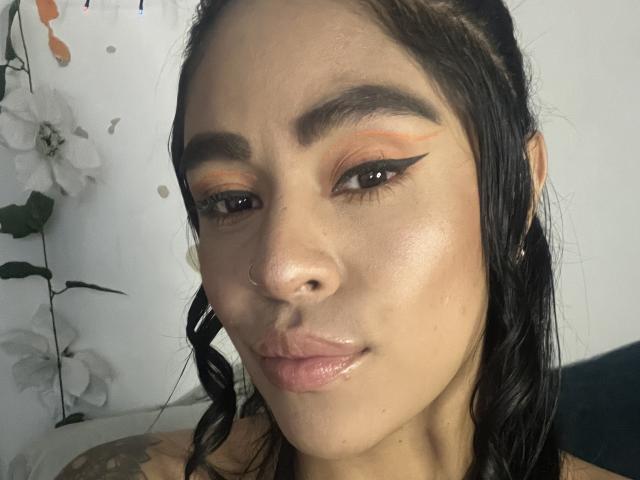 StellaBeckker has glossy, shiny lips and a nose ring. She's got dark, wavy hair, bold orange eyeshadow with black winged eyeliner, and a light, smooth skin tone.