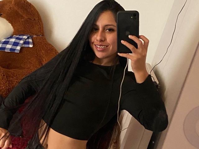 IsabelEvanss with long black hair, smiling, wearing a black crop top, holding a phone, mirror selfie, visible tan skin, small breasts, and a cute face, standing next to a teddy.