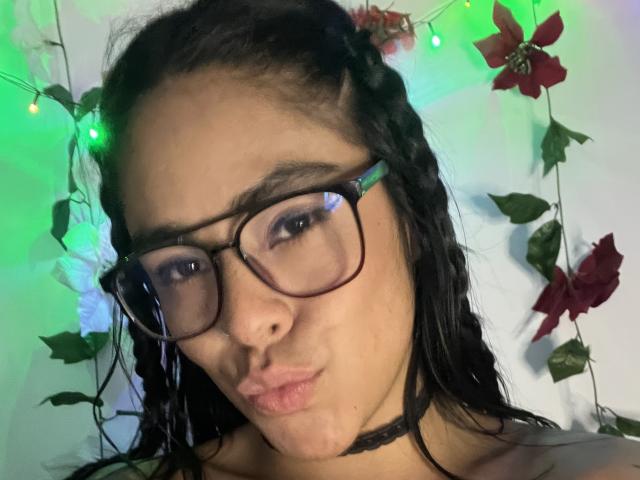StellaBeckker, com cabelo escuro em tranças, usando óculos e um colar preto, fazendo beicinho, cercada por luzes de Natal verdes e vermelhas e flores de Natal..